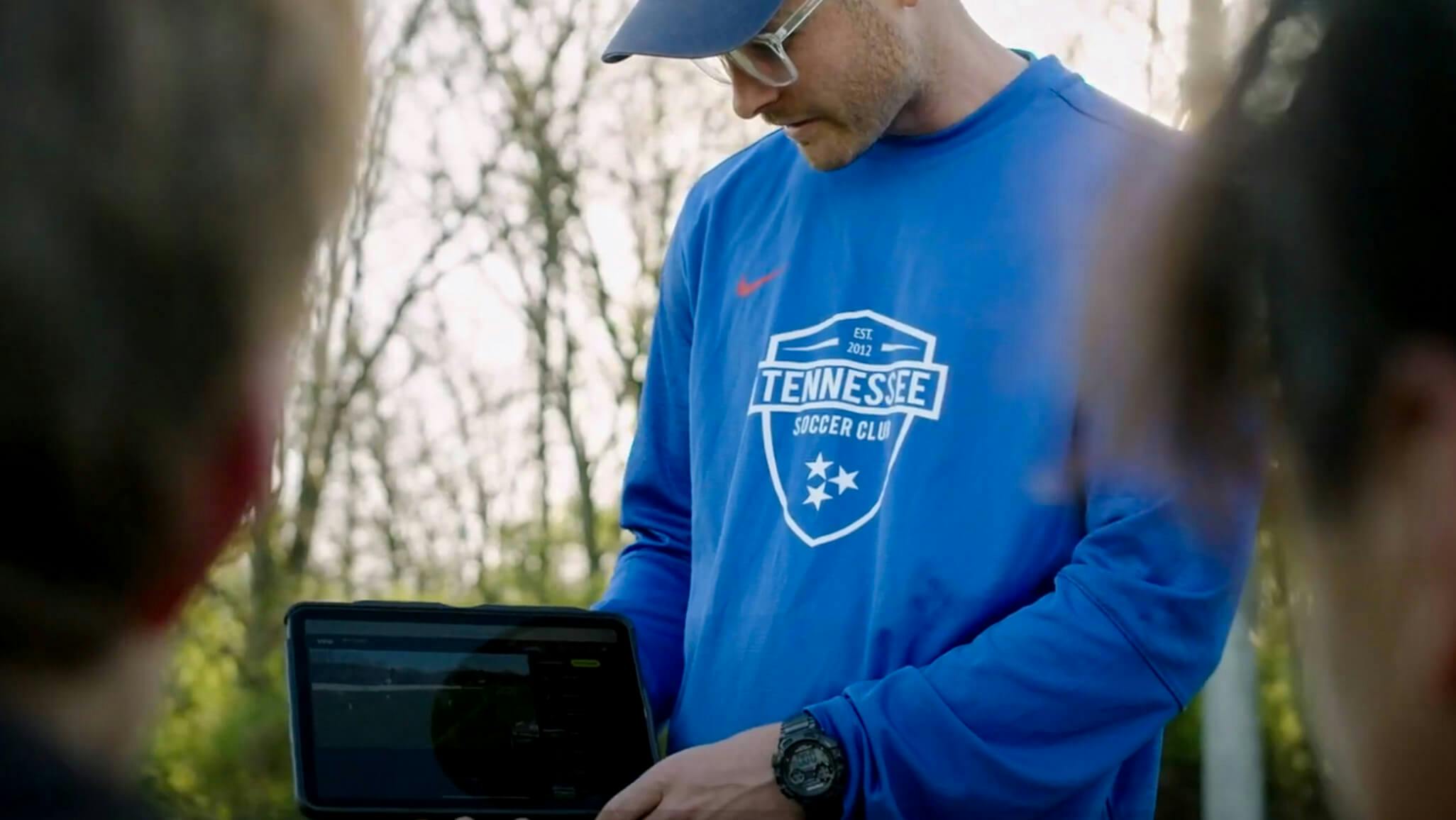 A photo of a coach from a Tennessee soccer club showing his players a video from their previous match recorded by a Veo sport camera, while they stand in a huddle on the soccer field.